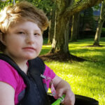 This is a photo of my daughter, Helen, that I took in 2019. She is sitting in her wheelchair on a sunny day, awaiting the schoolbus.