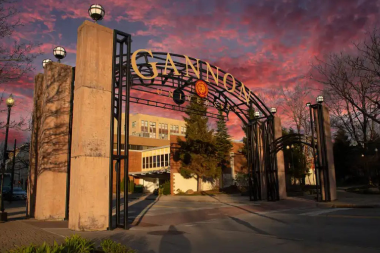 A photo of a beautiful arch bearing the name Gannon University in with flowers in front and a pink sky as the sun recedes.